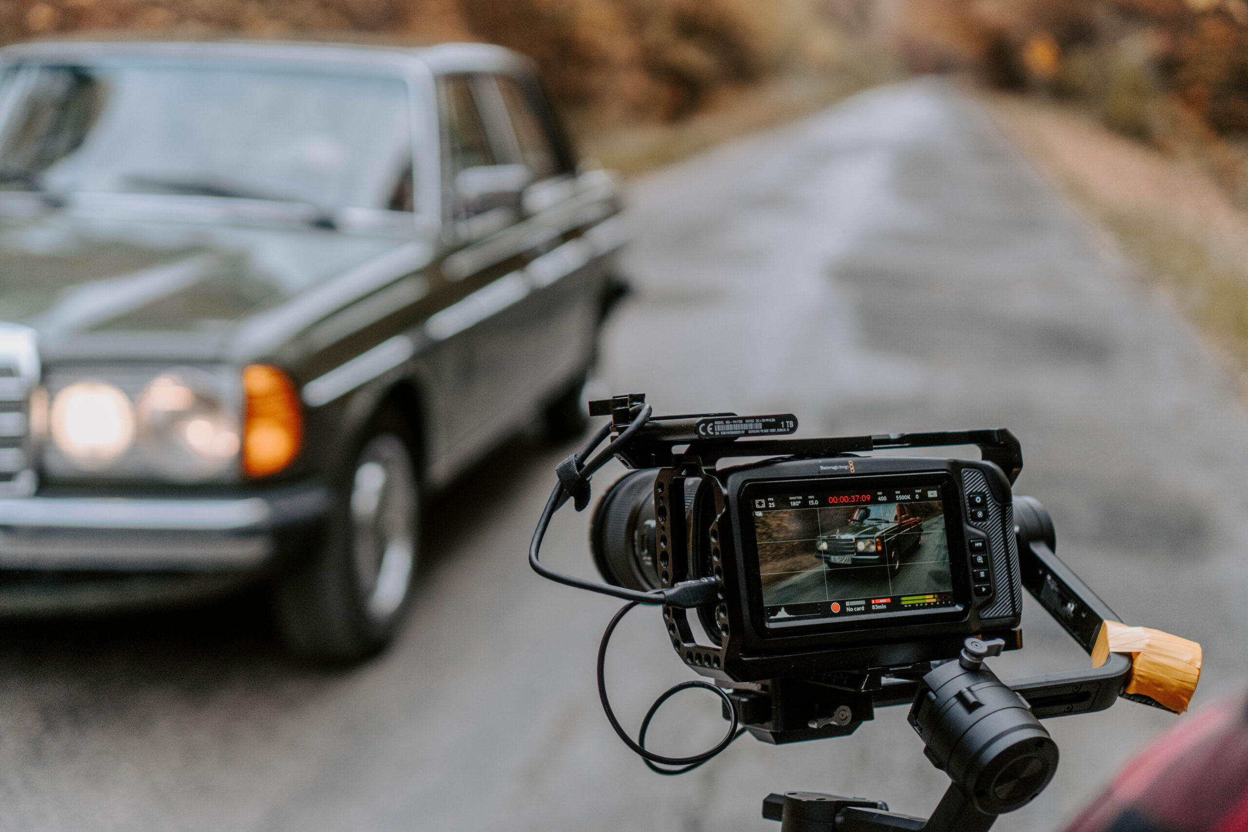 Zbliżenie na kamerę na stabilizatorze gimbal, nagrywającą klasyczny samochód na drodze. Na ekranie kamery widoczny jest obraz nagrywanego auta. W tle rozmazany widok asfaltowej drogi otoczonej drzewami.