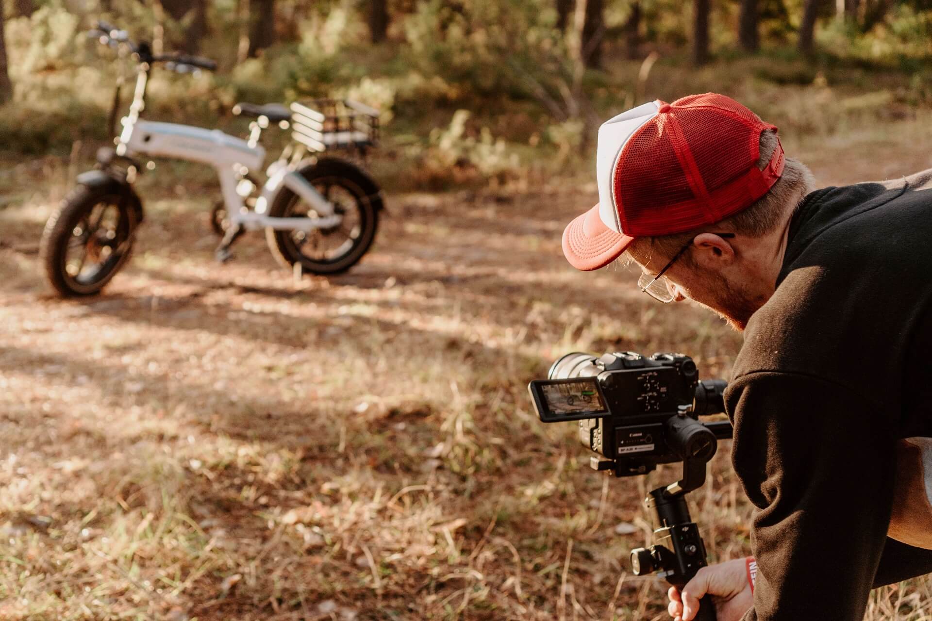 piotrek-creator Mężczyzna w czerwonej czapce z daszkiem i okularach, trzymający kamerę na stabilizatorze gimbal, nagrywający rower elektryczny na leśnej ścieżce w słoneczny dzień. Rower jest w tle, rozmazany, a mężczyzna skupia się na uzyskaniu idealnego ujęcia.