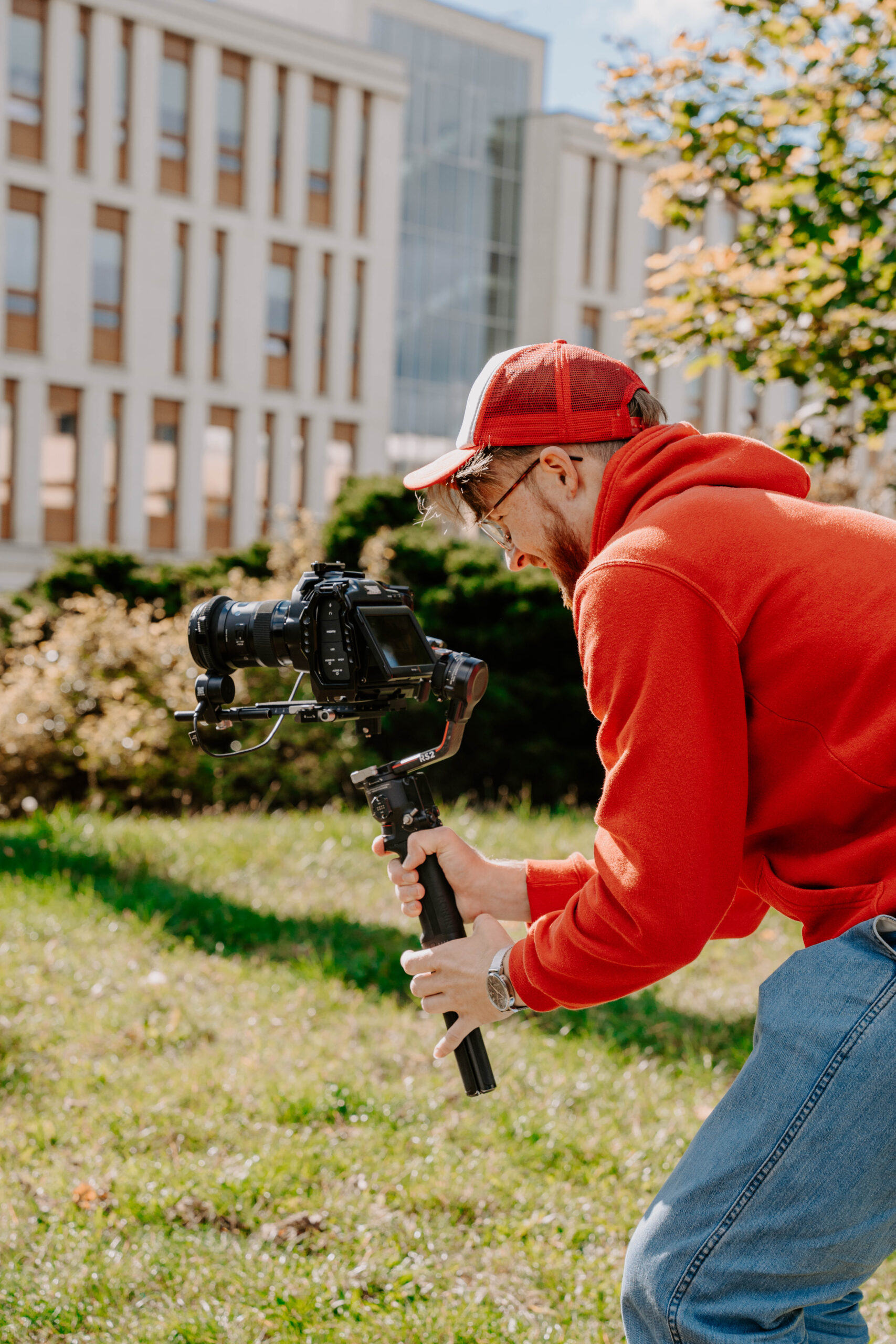Mężczyzna w czerwonej bluzie i czapce z daszkiem, trzymający kamerę na stabilizatorze gimbal, nagrywający na zewnątrz w słoneczny dzień, na tle nowoczesnego budynku z dużymi oknami i zielenią.